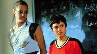 A tall, skinny man with long hair pulled back into two braids wearing a tight sleeveless shirt and small, round sunglasses, and a woman with short black hair in a red and black shirt stand in front of a chalkboard