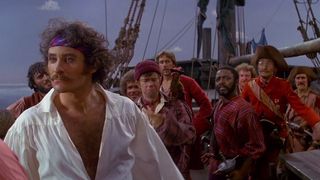 A mustachioed man in a puffy white shirt, open to his naval, and unruly hair held back by a colorful bandana stands on a ship in front of a crew of pirates looking seriously at someone just out of frame to the viewer’s left