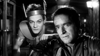 A black and white image of two men; one is wearing a strange crown/headband, a kind of armor-like vest over a bare chest, and a metallic arm band over his bare bicep as he leans on the shoulder of the other man in a leather jumpsuit while they both peer into the camera