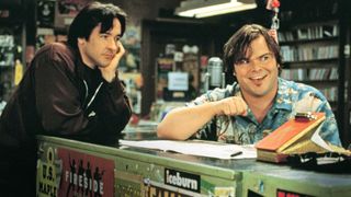 Two white men lean on a store counter, the front of which is covered in stickers; the man on the left is cupping his chin in his hands and looking at the man on the right, who is wearing a loud Hawaiian shirt and looking out of frame while grinning and making a fist-bump gesture; in the background we can see shelves containing what might be CDs or vinyl records