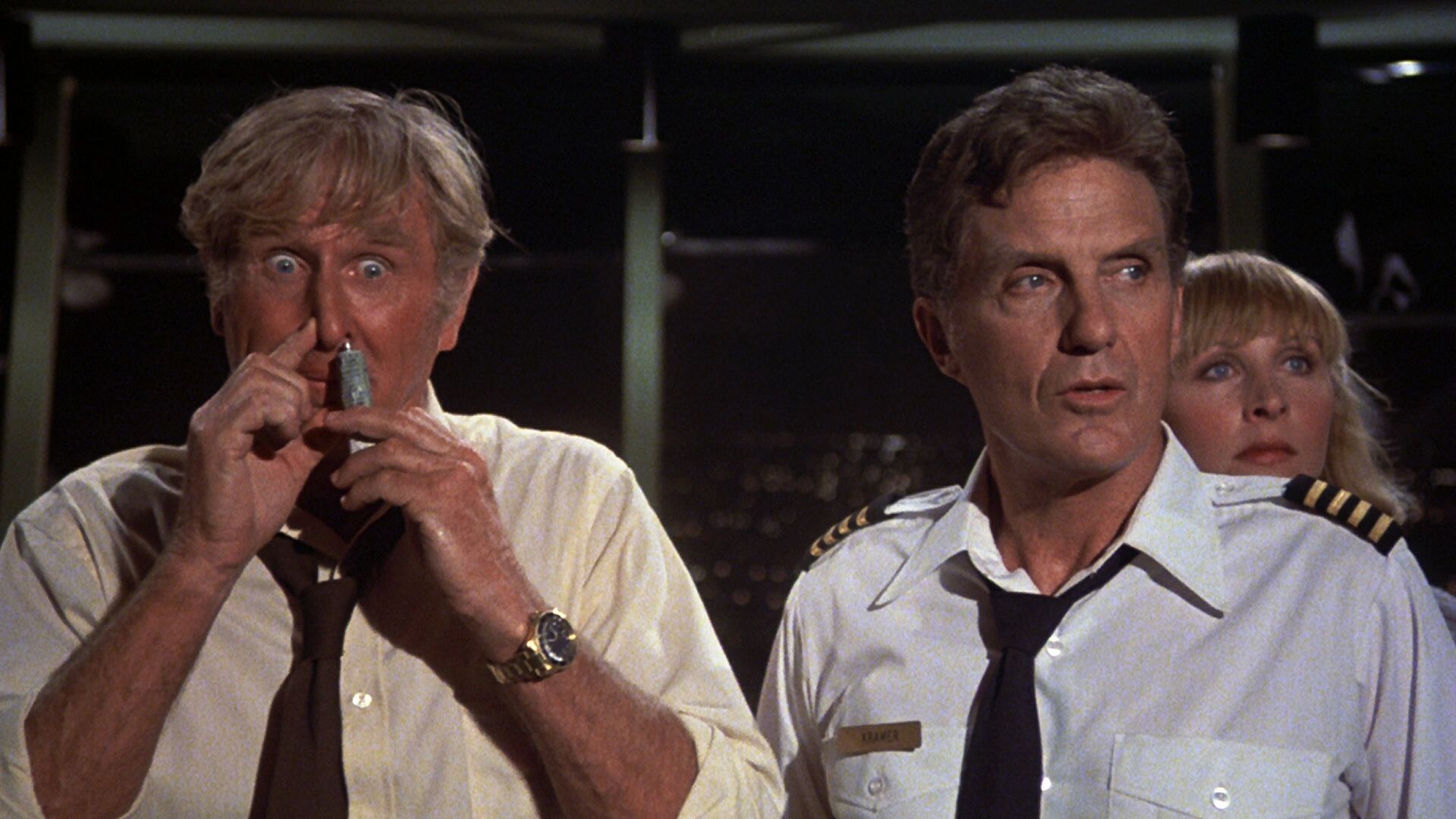 A wide-eyed man in light yellow shirt with a brown tie loose around his neck holds a tube of glue to one nostril while pressing the other nostril closed; to his left is another man in a pilot’s uniform, and just over his left shoulder we see the face of a blonde woman, both of whom are looking off to their left