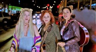 A blonde white woman, a red-headed white woman, and a brown haired black woman stand in the street, illuminated by headlights look wide-eyed with surprise into the camera; all three are wearing leopard print cat ears on their heads; the red-head has an acoustic guitar strapped across her front, and the brunette has an acoustic guitar strapped across her back