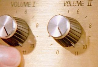 A close-up of two knobs labelled “VOLUME I” and “VOLUME II”, both of which go up to 11; the knobs are turned all the way up, and a finger points to the 11 of the first volume knob