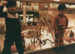 Two young kids pose in front of a collection of dinosaur skeletons