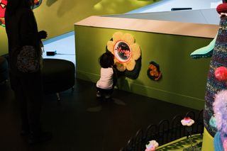 A kid crouches in front of a mirror framed with crotcheted yellow flower petals, written on the mirror are the words “you are strong”