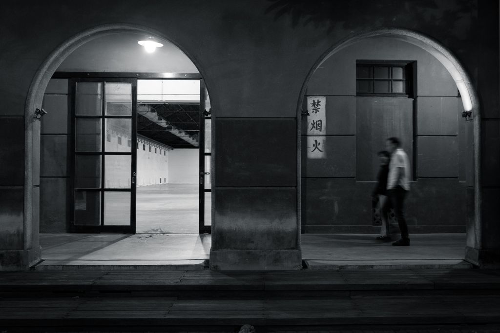 A black and white photo of two arches of a cloister; in the left arch, a lamp hangs from the ceiling, illuminating a double sliding glass door leading to a large empty room with white walls, a concrete floor, and an open ceiling with visible duct work; in the right arch two blurry figures in the center of the arch are walking along the cloister towards the left edge of the frame, the wall behind them is dark except for a vertical white rectangle containing three characters written in Chinese
