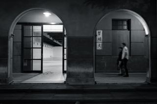 A black and white photo of two arches of a cloister; in the left arch, a lamp hangs from the ceiling, illuminating a double sliding glass door leading to a large empty room with white walls, a concrete floor, and an open ceiling with visible duct work; in the right arch two blurry figures in the center of the arch are walking along the cloister towards the left edge of the frame, the wall behind them is dark except for a vertical white rectangle containing three characters written in Chinese