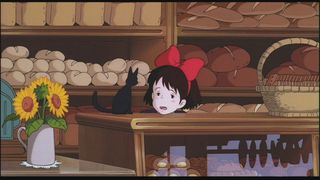 A frame from an animated film where a young, tired-looking girl rests her chin on the counter of a bakery in front of shelves piled high with bread; she has shoulder-length brown hair, bangs, and a large red bow holding her hair back, and she is looking at a small black cat who is sitting on the counter turned away from the camera