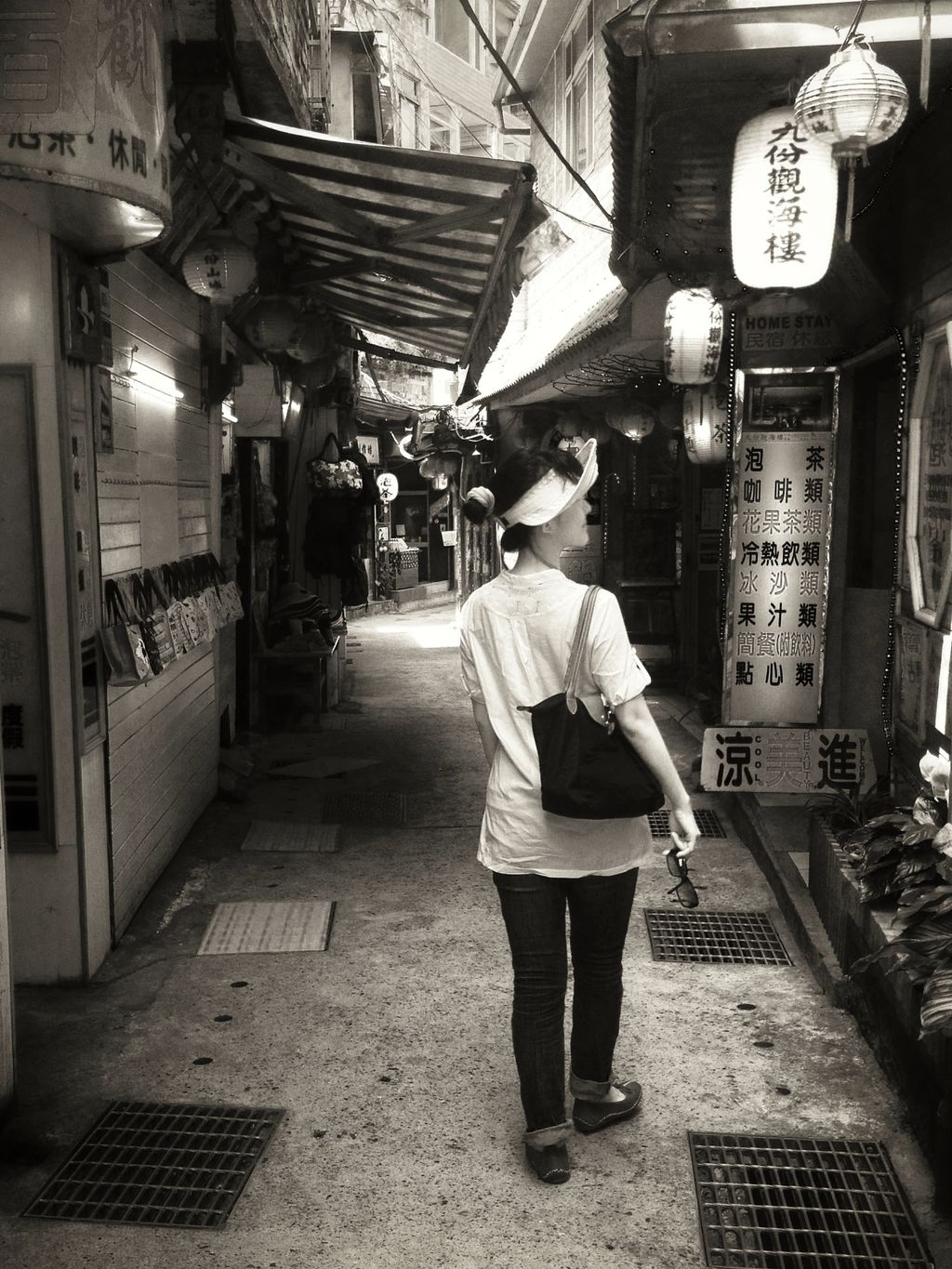 A black and white photo of a woman in a white shirt and dark trousers with a purse hung over her right shoulder walking down a narrow street lined with shops; most of the shops have paper lanterns of various shapes and sizes with Mandarin characters written on them hung outside
