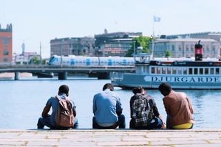 Four people sit on a concrete curb on the bank of a river with their backs to the camera on a sunny day; in the background, a boat on the river protrudes from the right side of the frame, and a train is visible crossing a bridge behind it