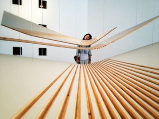 Three panels of plywood hang horizontally suspended from wires; the panels are each bent to different degrees and hung from different heights, they have grooves cut into them; behond the plywood a woman with long dark hair stands with her head tilted to the right to match the curve of the plywood in front of her as if she were resting her head on it