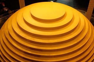 An enormous tiered yellow sphere lit from above; the sphere is made of short, progressively smaller cylinders stacked atop one another