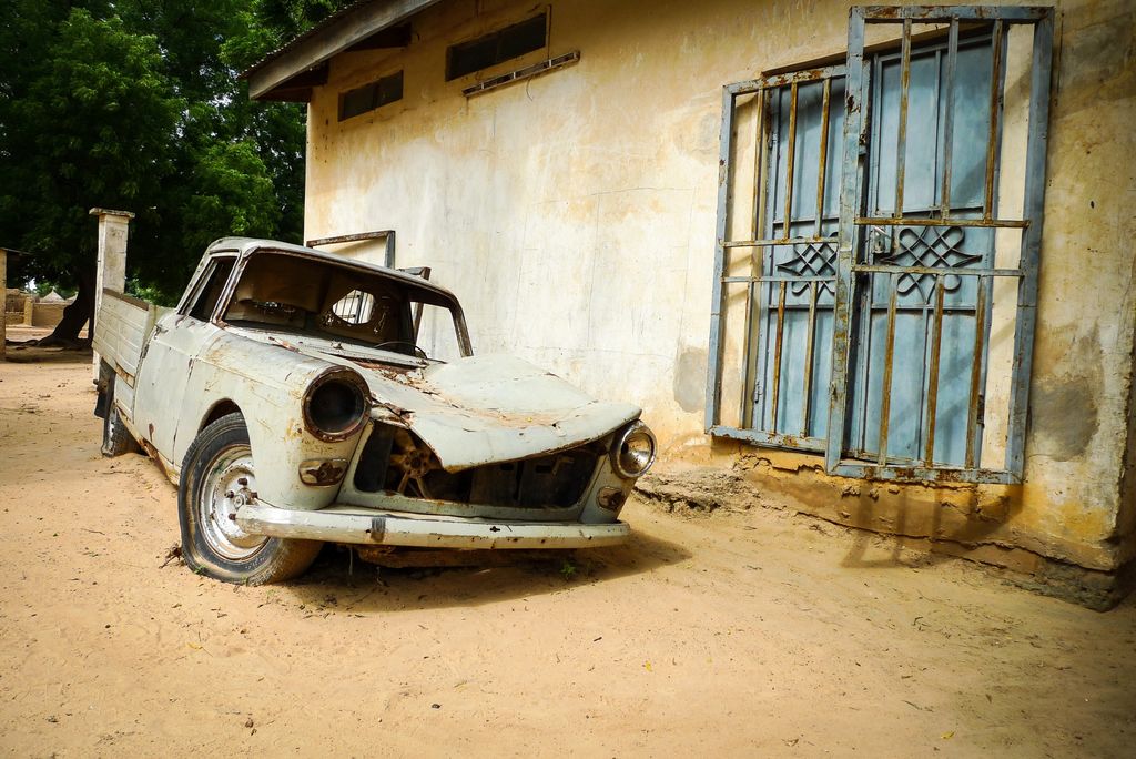 A broken down truck with a flat tires, no windows, a missing headlight, and a smashed hood sits in yellow dirt in front of a plain building; the truck is listing toward the driver-side front wheel as if that wheel is missing
