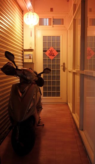 A narrow hallway leading to a door with a piece of square red paper taped to it; the square is rotated 45 degrees so that the corners of the square are oriented vertically and horizontally, and on the square is a single, complex piece of Chinese calligraphy; the hall is illuminated by a red paper lantern with a tassle hanging from the bottom hanging just in front of the door on the left of the frame, and a scooter is parked in the hall, taking up about one third of the hall on the left side of the frame in front of a metal garage door