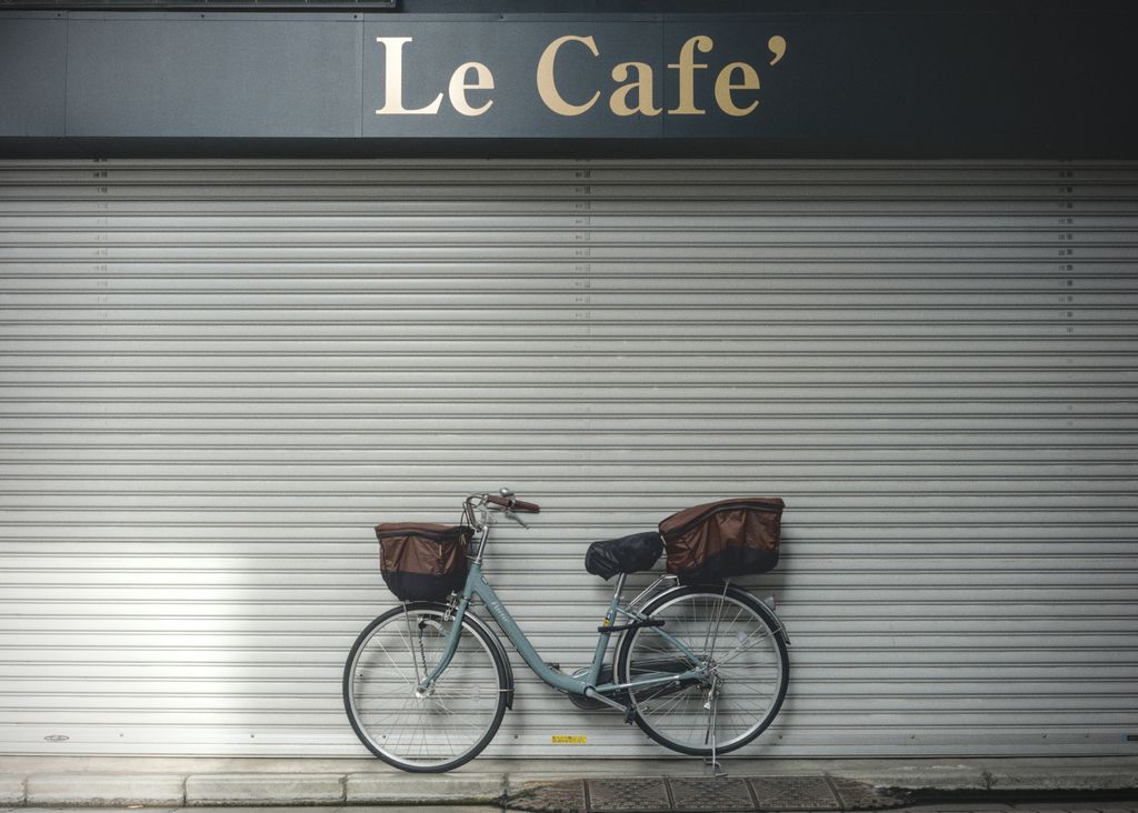 A teal step-through bicycle with bags front and back parked in front of a metal garage door of a closed shop under a sign that reads “Le Cafe’”