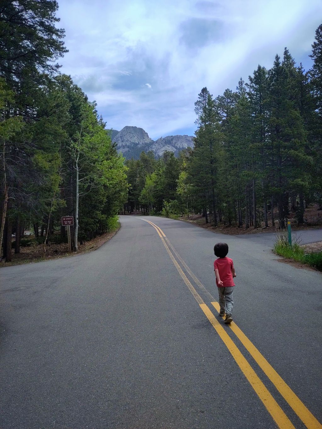 A young kid in a red shirt walks down the middle of a two-lane road swinging his arms, the road is lined with aspen and evergreen trees, in the distance is a rocky mountain peak