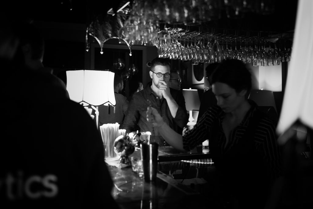 A black and white photo of a man in the middle distance standing at a bar; he is holding his hand over his mouth, with one finger under his nose and the other three curled around his chin, and is wearing round glasses, a dark button-down shirt, and has short, dark hair and a close-cropped beard; he is illuminted from the left side of the frame by an old table lamp with tassles hanging from the shade; dozens of wine glasses hang by their stems from the ceiling, and in the foreground a woman bartender in a dark shirt with white vertical stripes mixes a drink