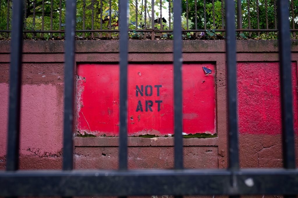 Seen through the bars of a black wrought iron gate, the words “NOT ART” in all caps have been spray painted in black through a stencil onto a bright red patch of wall, each word is on a line by itself so that the letters of each word line up with one another; the surrounding wall is a dull brown with patches of red paint; at the top of the wall is more wrought iron fence, behind which is lush greenery