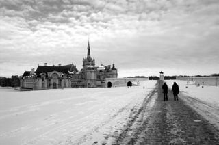 A black and white photo; in the background, in the left half of the frame, a large chateau including a tall spire with a cross atop is surrounded by a field of snow; on the right half of the frame two people in winter coats walk away from the camera down a road paved with cobble stones; at the end of the road, in the distance, is a large statue of a person riding a horse; the sky is covered with clouds