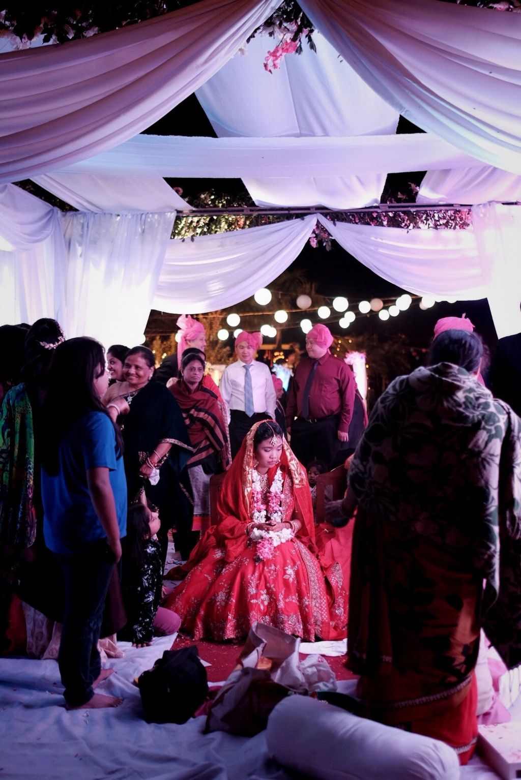 A young woman in an elaborate red dress with a necklace of flowers sits underneath a canopy of white fabric at night among a crowd of people in suits and dresses, some of whom are wearing bright pink turbins; she is facing the camera with her head turned down and to the right