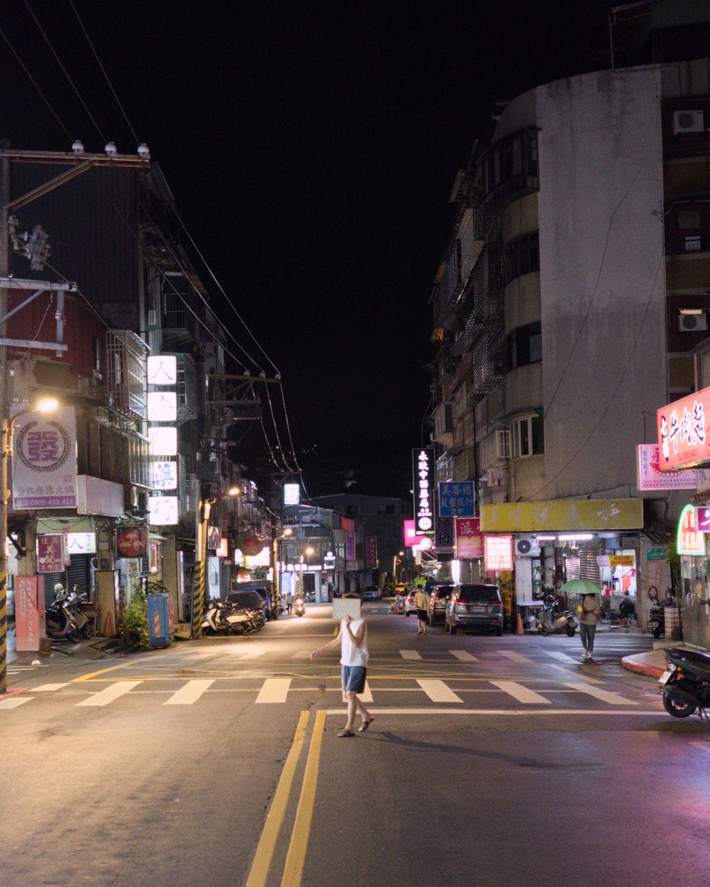 A person crosses a quiet, rain-wet street at night, holding up a piece of paper to cover their face; the street is lit by yellow street lamps and colored shop signs