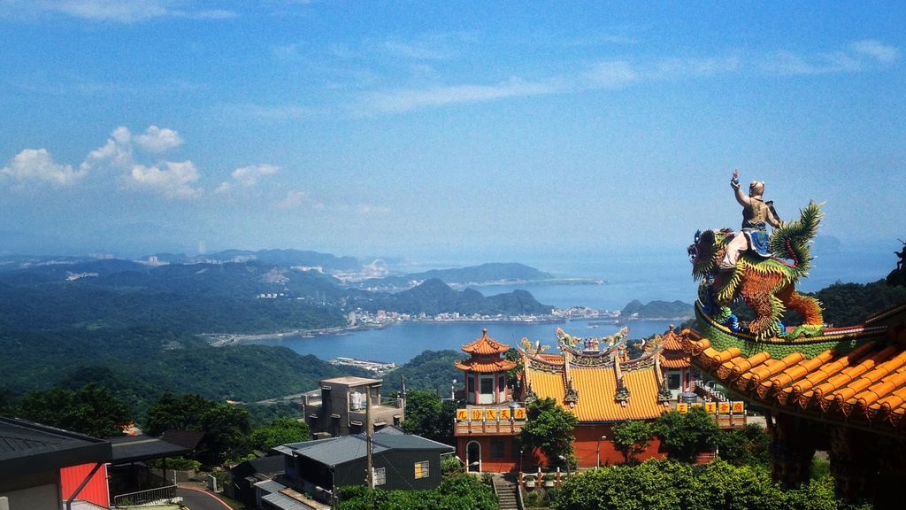 At the right of the frame in the foreground is the corner of a roof covered with round, bright yellow tiles; atop the corner, facing away from the camera toward the center of the frame, is a statue of a person with their left arm raised riding some kind of monster whose green, yellow, and orange fur resembles a sea anemone more than actual fur; the statue looks out over a bay surrounded by tree-covered mountains; a smattering of buildings is just visible along the coast