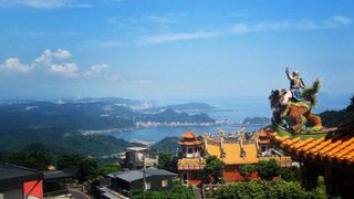 At the right of the frame in the foreground is the corner of a roof covered with round, bright yellow tiles; atop the corner, facing away from the camera toward the center of the frame, is a statue of a person with their left arm raised riding some kind of monster whose green, yellow, and orange fur resembles a sea anemone more than actual fur; the statue looks out over a bay surrounded by tree-covered mountains; a smattering of buildings is just visible along the coast