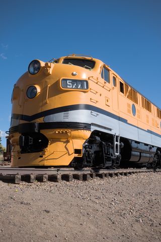 A bright yellow diesel train engine with a thick, horizontal gray stripe, and a thinner horizontal black stripe running down the length of the engine just above its wheels; the front of the engine is rounded and has two large headlights, one above the other, on the front; the number 5771 is visible on one side of the lower headlight