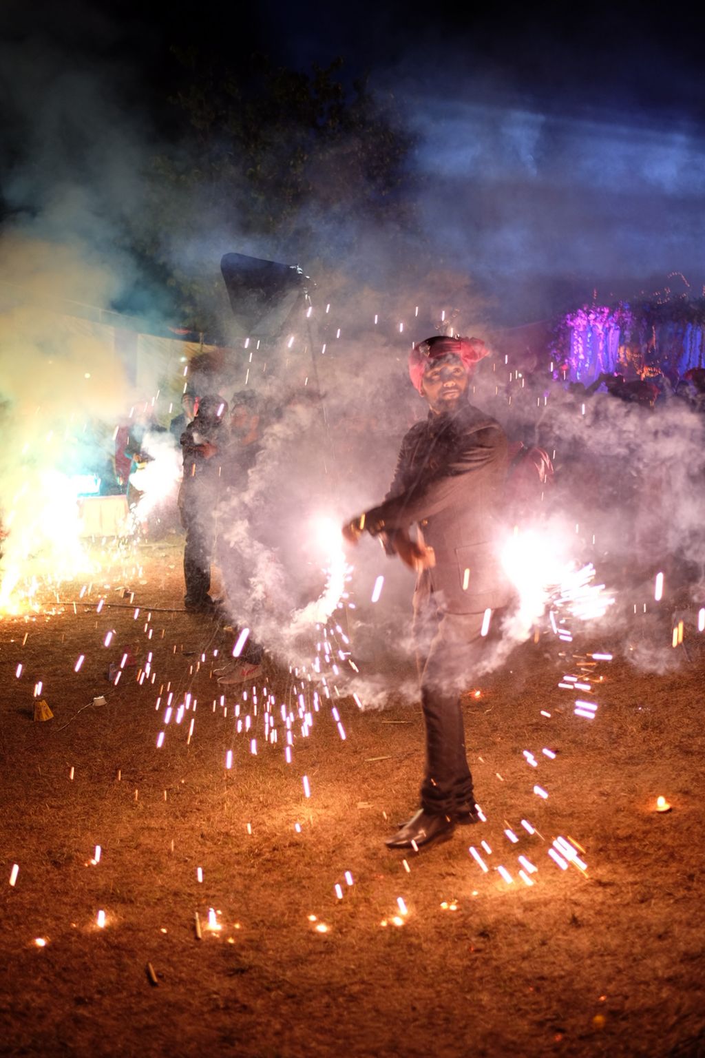A man stands on a field of short, dry grass in a dark suit and bright pink turbin twirls two sparklers around him with his arms crossed in front of his body as he looks off into the middle distance towards the right side of the frame; he is surrounded from the knees up by smoke from the sparklers; just visible the the haze of the smoke, a crowd of people mills around behind him, with more smoke and sparks visible from other fireworks