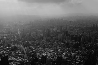 A black and white photo of a dense city pocked with skyscrapers; rays of sun shine through hazy clouds over the city, and in the distance sillhouettes that hint at the presence of mountains