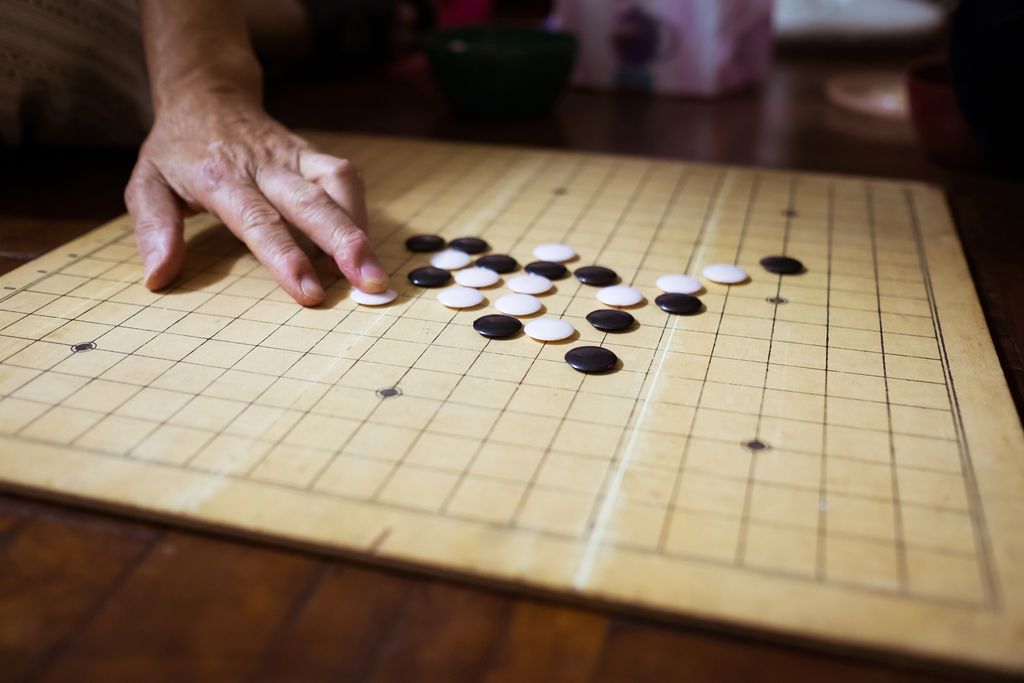 A Go board sits on a wood floor with two dozen black and white pieces on it; a person’s hand is visible on the left side of the frame with their middle finger resting on a white piece after having just placed it on the board