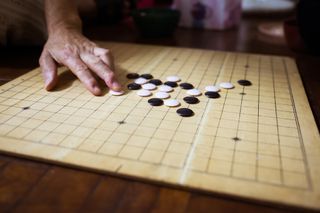 A Go board sits on a wood floor with two dozen black and white pieces on it; a person’s hand is visible on the left side of the frame with their middle finger resting on a white piece after having just placed it on the board