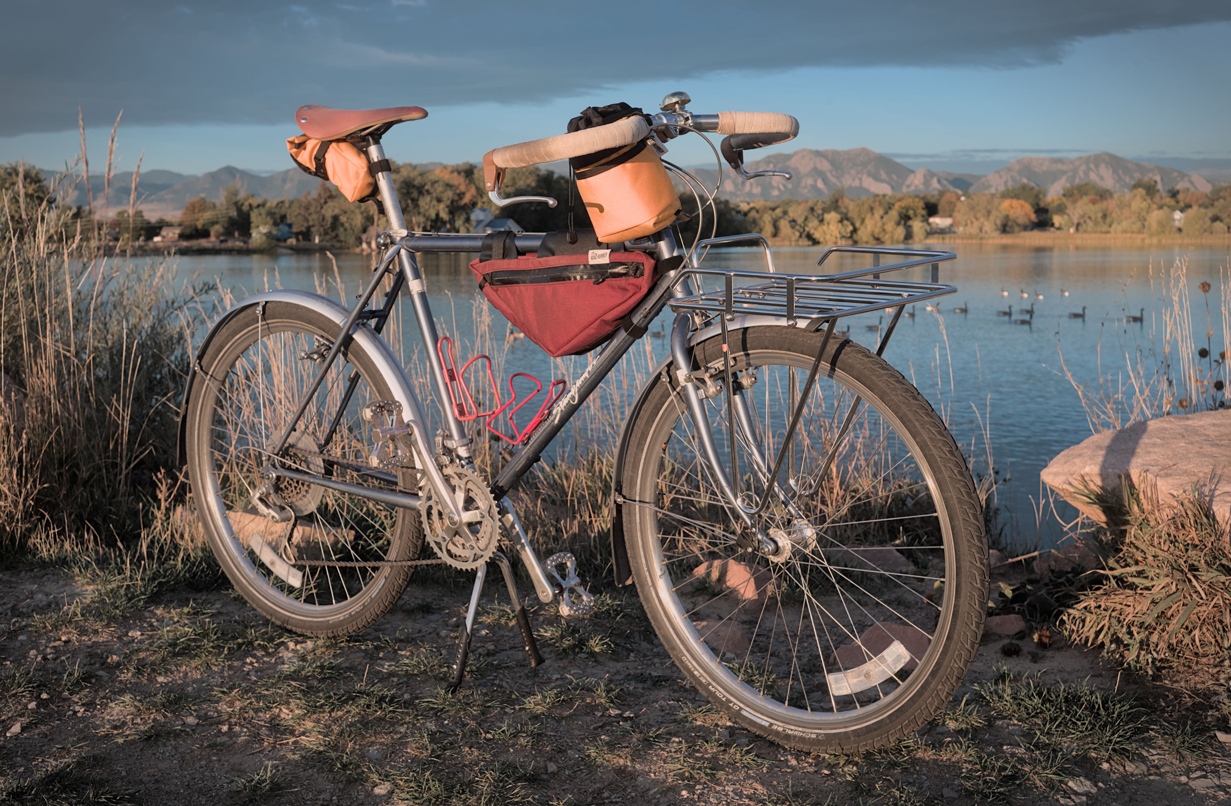 A blue bike with a chrome front rack and several frame bags parked in front of a lake with a mountain range in the background