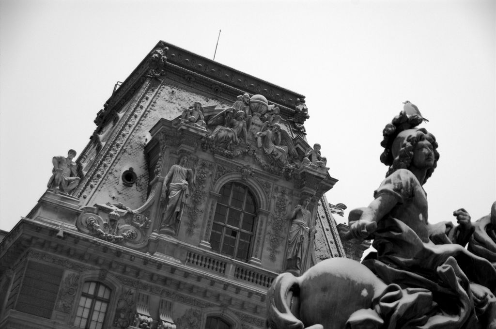 A black and white photo of an ornate building taken at a jaunty angle; the visible portion of the building forms a trapezoid, it has a large window in the middle that is surrounded by ornate sculptures of people and flowers; in the foreground at the bottom right of the frame, a portion of a statue of a person riding a horse is visible; a real bird sits atop the statues head; both the building and the statue are covered with a light dusting of snow