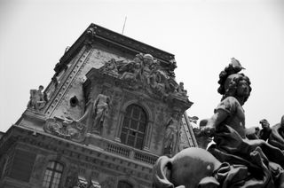 A black and white photo of an ornate building taken at a jaunty angle; the visible portion of the building forms a trapezoid, it has a large window in the middle that is surrounded by ornate sculptures of people and flowers; in the foreground at the bottom right of the frame, a portion of a statue of a person riding a horse is visible; a real bird sits atop the statues head; both the building and the statue are covered with a light dusting of snow