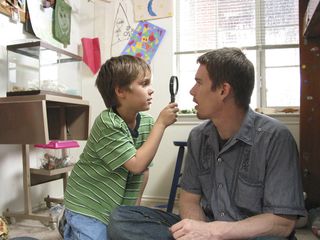 A young boy kneels on the floor next to an adult man who sits cross-legged on the floor of a room with kid’s art on the walls; the boy holds a magnifying glass up to the man’s face