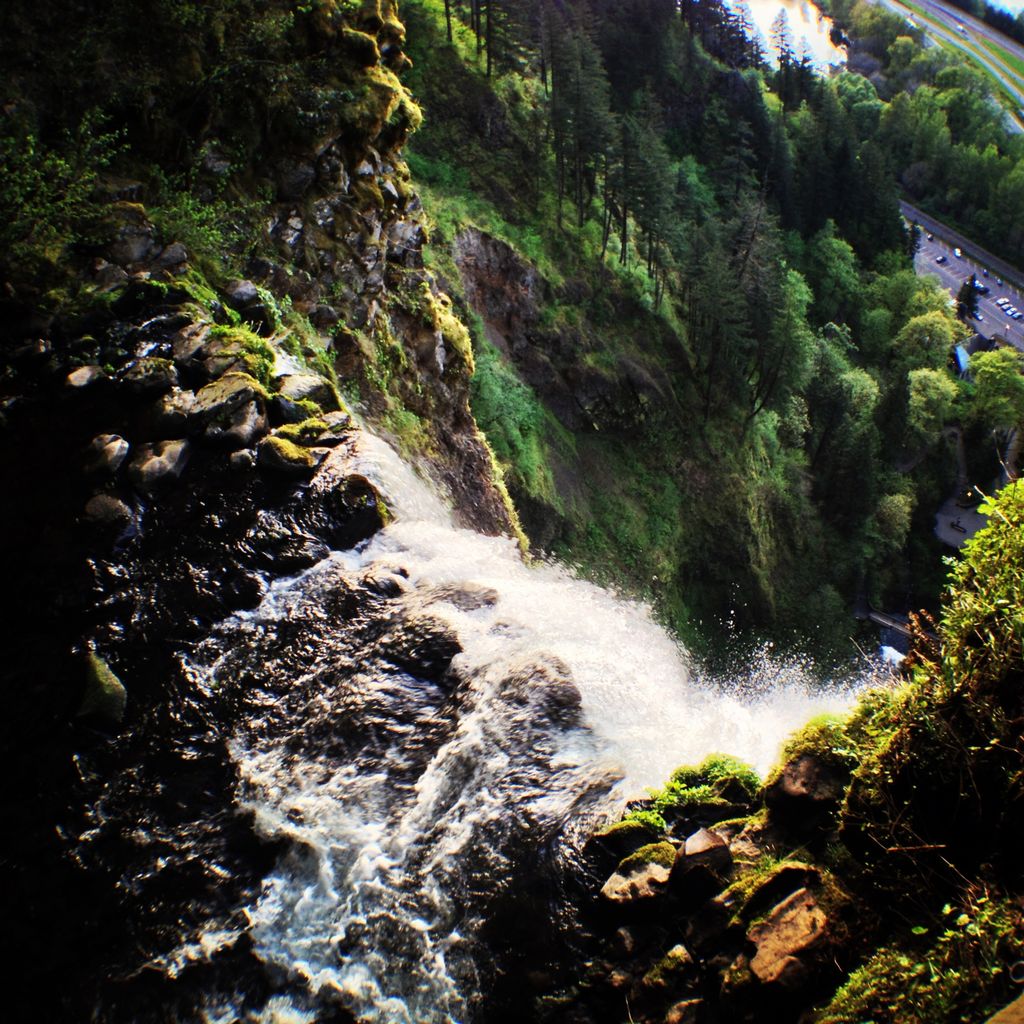 Water cascades over an abrupt cliff’s edge, sharp rocks covered with plant life frame the falls; in the distance, steep mountainsides covered with plants and trees give the impression that the photo was taken from high up