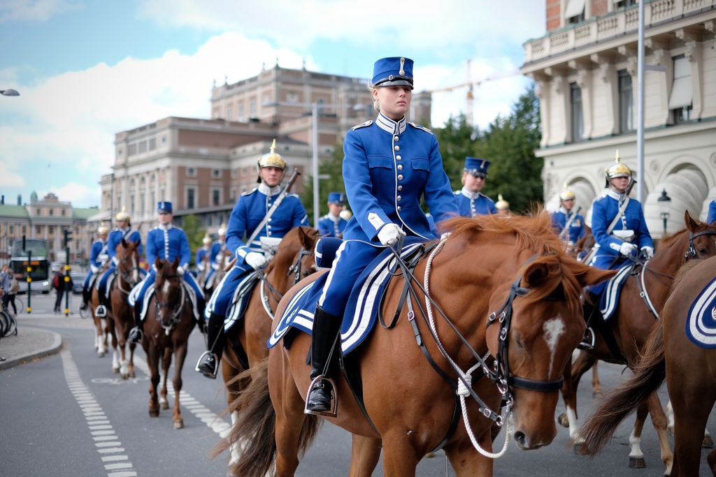 A parade of people in bright blue uniforms, some wearing cylindrical hats, others with silver and gold helmets with a spike on top, ride horses down the street toward the camera