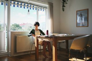 A young asian woman in a white, knee-length dress and a blue and green striped cardigan sits in a white, plastic, modern chair at a wooden kitchen table and looks into the camera with a serious expression on her face; her hair is cut short—down to the bottoms of her ears—and frames her eyes as she looks into the camera with a serious expression on her face; behind her is a large window and a partially opened patio door; treetops and a red roof are visible through the window