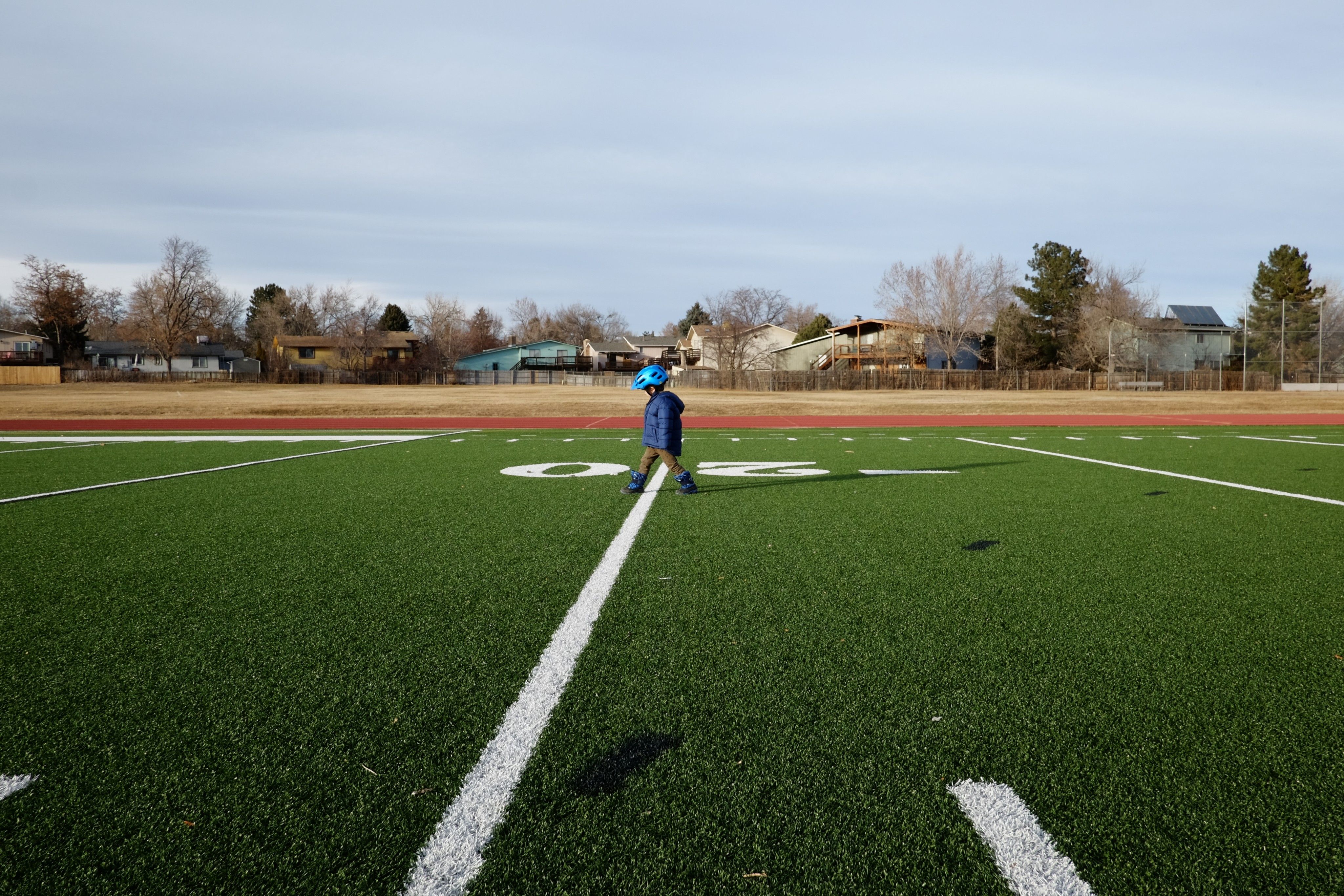 A toddler in the center of the frame wearing a blue puffy jacket and a bright blue bike helmet steps over the 20 yard line as he takes long strides across a green football field; in the distance a number of single family homes with bare trees is visible on the other side of a field of yellowish brown grass, in stark contrast to the bright green of the football field