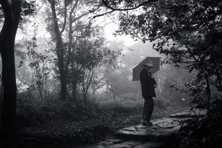 A black and white of a woman holding an open umbrella on a foggy day standing on a stone path; she is wearing a light colored hat with a brim that goes all the way around, a dark jacket, and dark trousers and is carrying the umbrella over her shoulder as she looks down at the ground towards the right edge of the frame; the path is paved with irregularly cut stones that are placed to form a relatively flat path with shallow steps leading from teh bottom center of the frame off to the right edge of the frame and is surrounded by trees and bushes