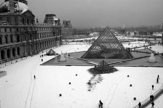 A black and white photo of a large courtyard covered in snow with a a few people walking around; the courtyard is dominated by a large glass pyramid surrounded by fountains; two smaller pyramids are placed just outside fountains surrounding the larger pyramid; the building forming one edge of the courtyard is old and ornate, covered with many statues and lined with arches along the ground
