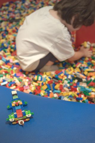 A Lego fighter jet sits on a low blue wall surrounding a pool of Lego pieces, in the background a child crouches with his back to the camera intent on building something