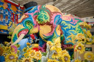 A psychadelic, multi-color Lego sculpture of an elephant in a top hat surrounded by (real) sunflowers