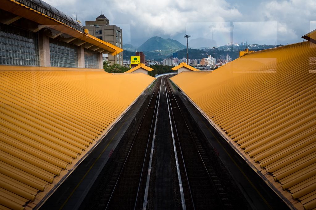 Two sets of train tracks run down the center of the frame and then veer off to the right, they are surrounding by bright yellow roofs on the left and right; the roof on the left ends in the wall of a second story made of tiled glass blocks topped with a similar yellow roof; in the distance are tall buildings, and beyond that green mountains