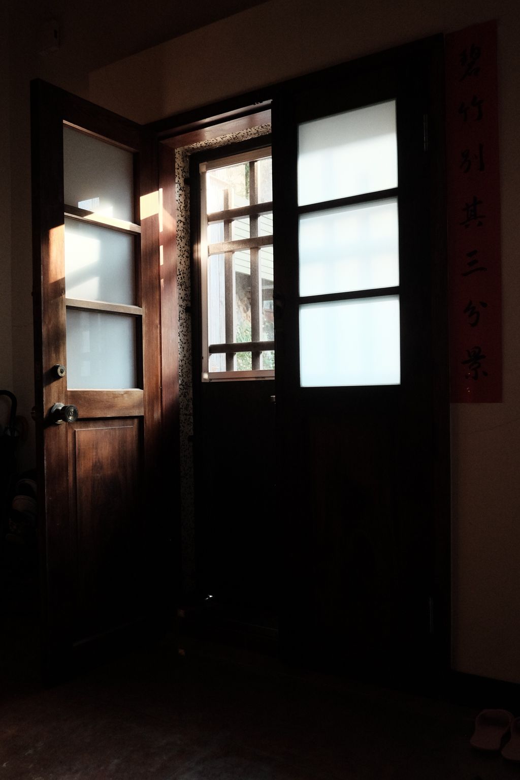 One half of a set of old, wooden double doors stands open, afternoon light streaks across the open door and the three panels of opaque white glass set in its top half, leaving the bottom half of the door in shadow; another outer door, this one closed, with thick metal bars is visible through the open door; the other half of the double-doors is visible only as three white rectangles of the glass set in the top half