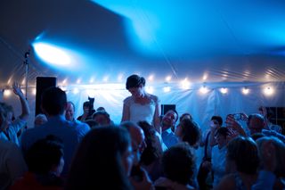 A woman in a white gown laughs as she is lifted up by a crowd of people inside a large white tent; the tent is illuminated by string lights and camera flashes
