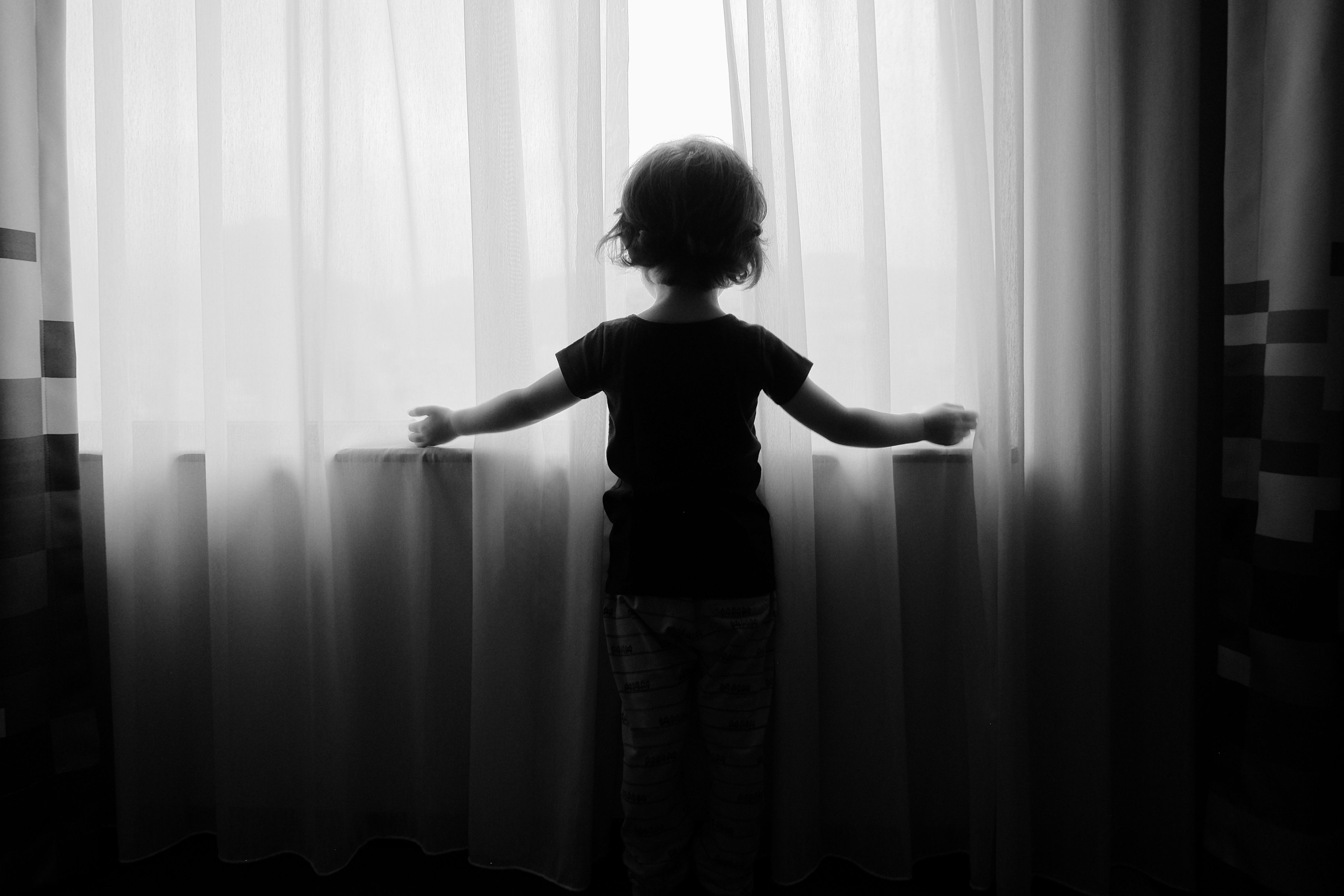 A black and white photo of a toddler standing at a window with his back to the camera looking out between a pair of sheer curtains with his arms spread wide to either side; the light coming in through the window is so bright that the window appears to be a solid white wall, nothing on the outside is visible