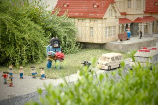 A Lego sculpture of a large, blue ogre holding a spiked club tromping around a city street with Lego people (not mini-figs), Lego cars, and Lego houses