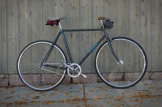 A dark framed, fixed gear bike leans against a wood wall, the sidewalk is strewn with leaves
